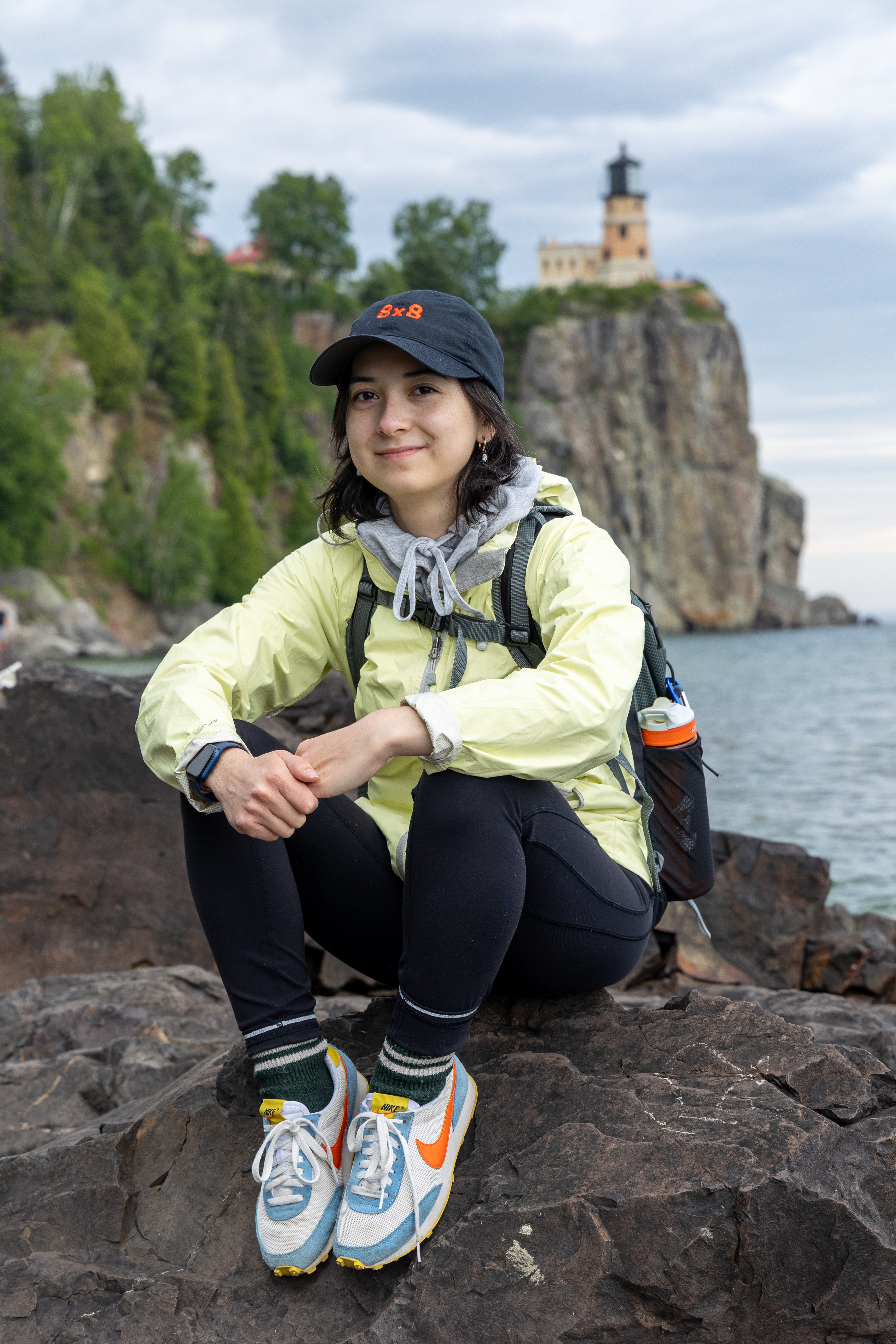 Portrait of Olive Phan in hiking attire sitting on a rocky surface in front of the Split Rock Lighthouse in Duluth Minnesota